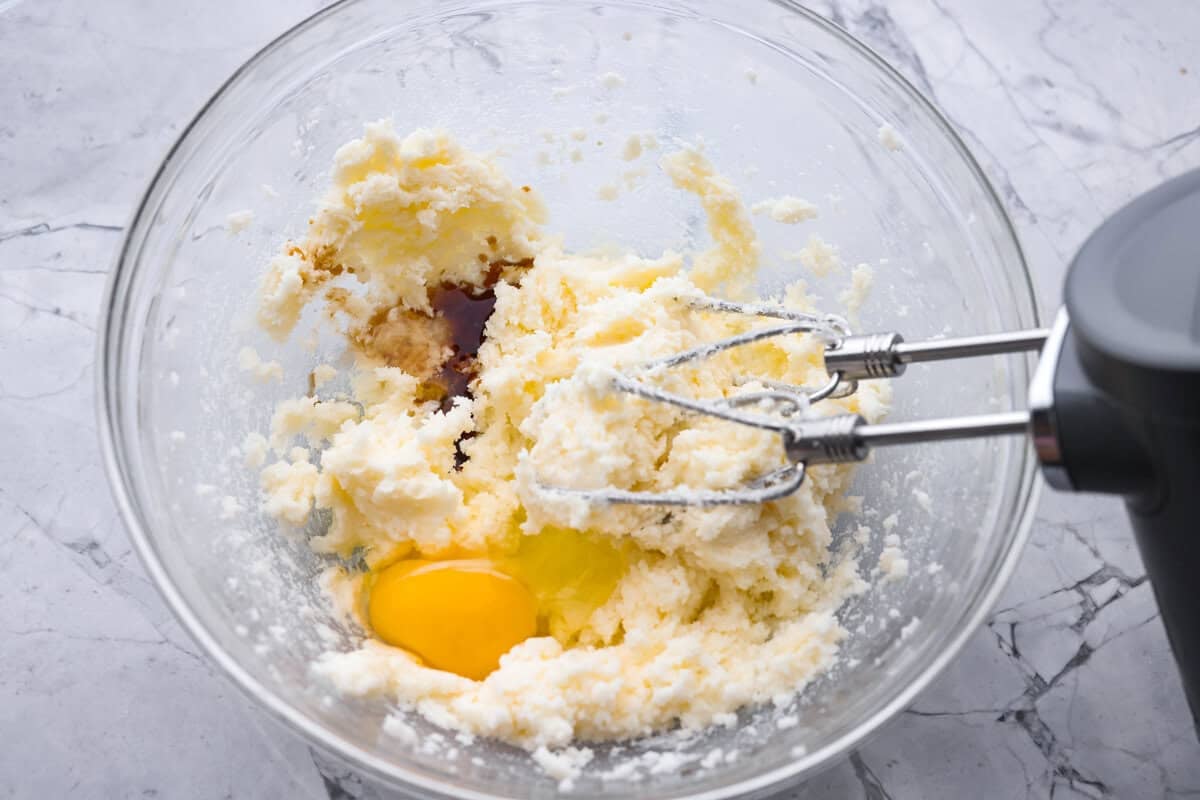 Someone using a hand mixer to beat the butter and sugar together. and an egg and extract added to the beaten butter and sugar.