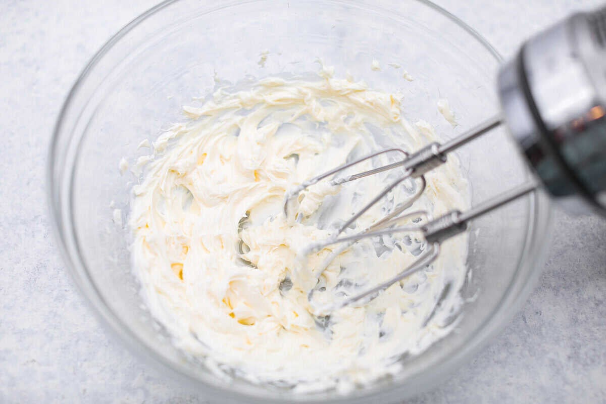 Cream cheese being beaten with a hand mixer in a mixing bowl.