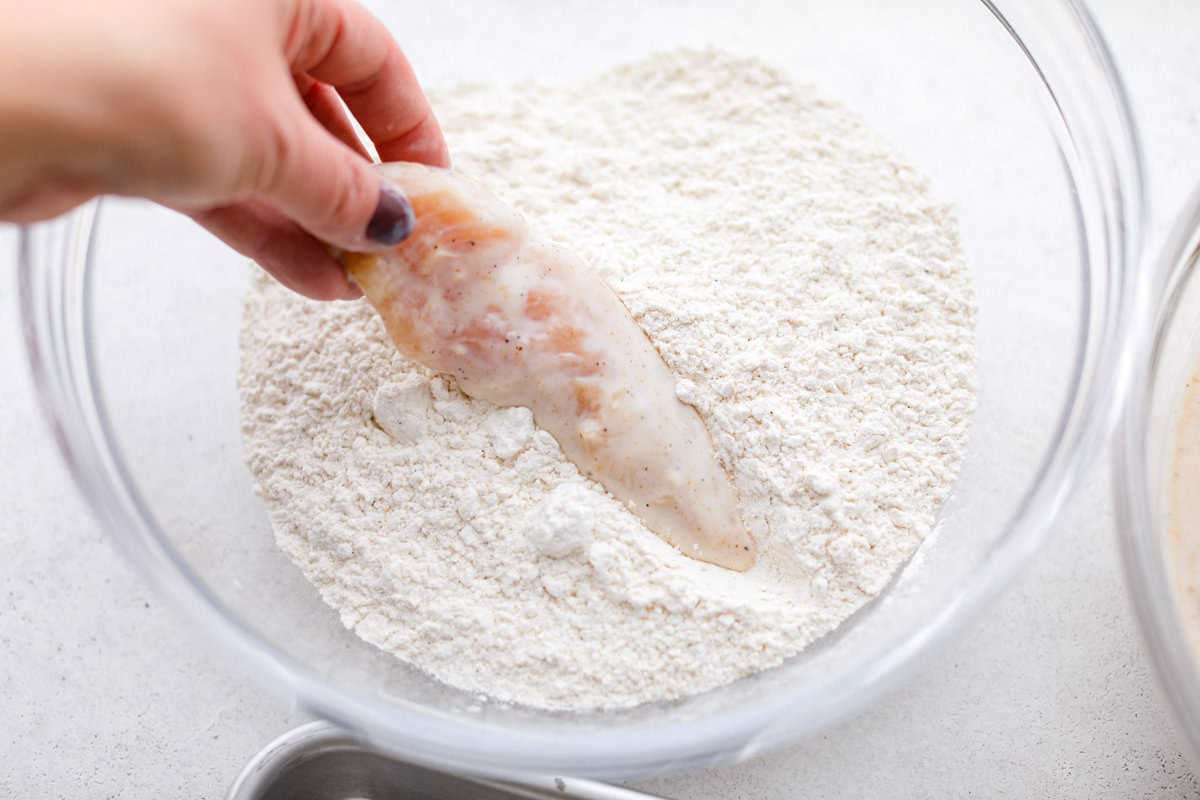 Someone dipping a marinated chicken tender into the bowl of breading ingredients.